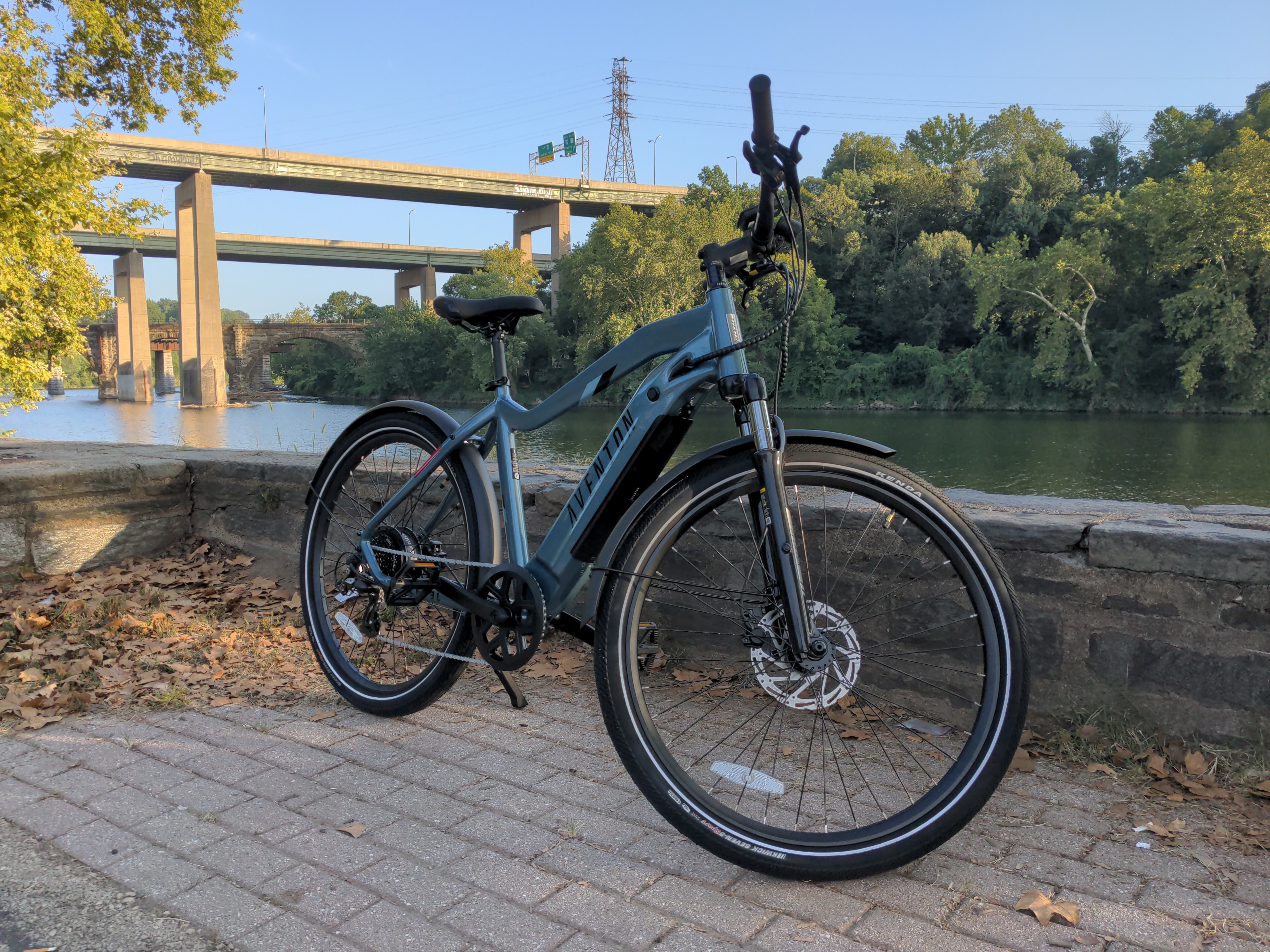 Photograph of a blue ebike in front of a river, with trees and bridges in the background.