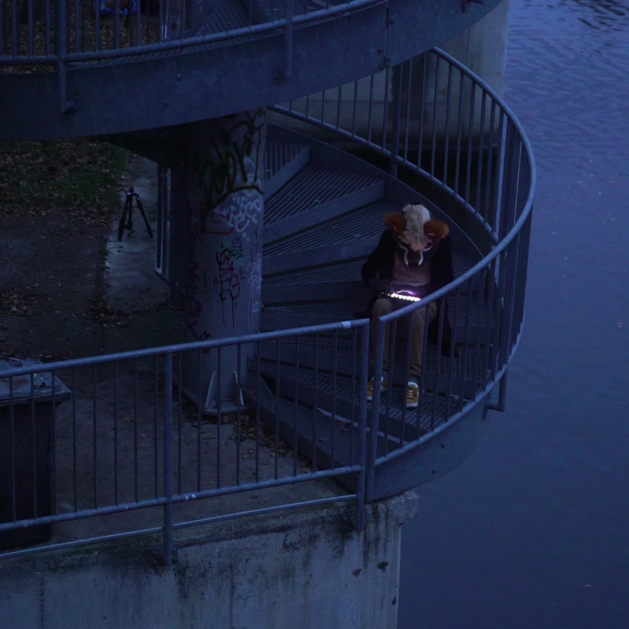 A person with a furry head sits on a circular metal staircase near a body of water and operates a glowing tablet.