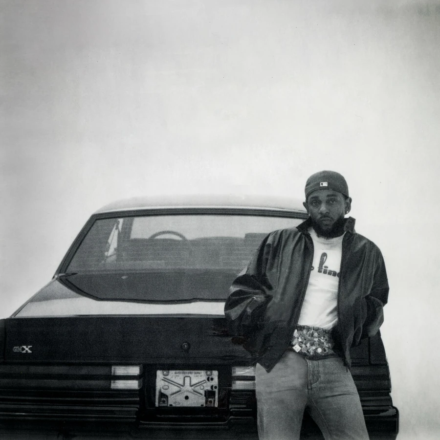 Kendrick Lamar, wearing a black jacket and blue jeans, stands in front of a black car.