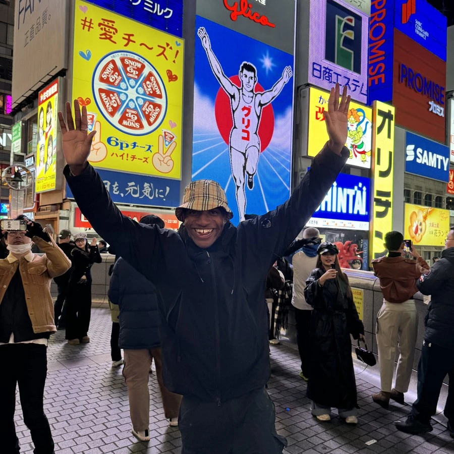 A man smiling and posing as if he is running with his hands raised, in the middle of a crowded street in Tokyo.